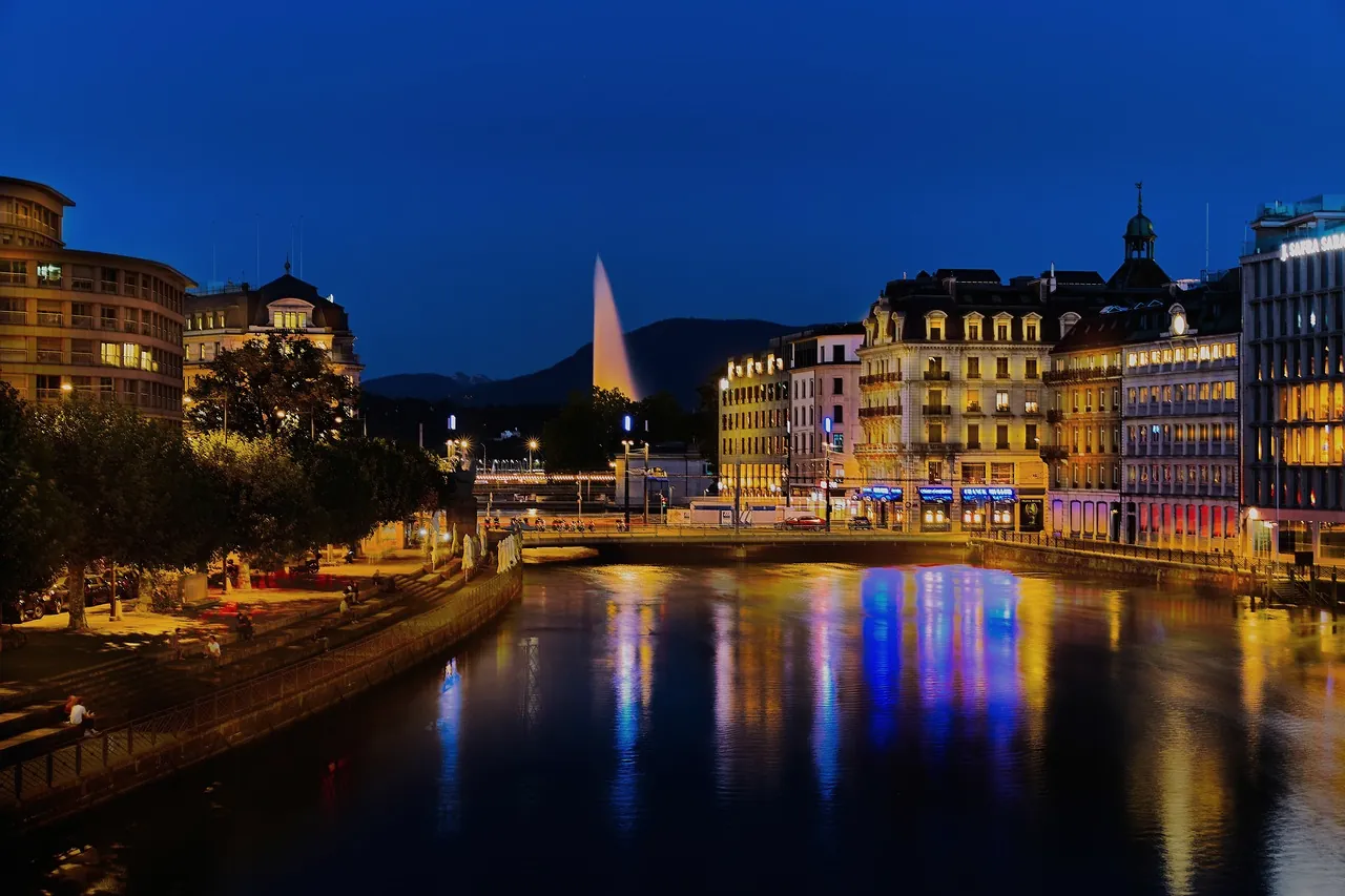 Geneva at blue hour: lit hotel facades along the Rhone, their reflections in the water and the Jet d'Eau in the distance.