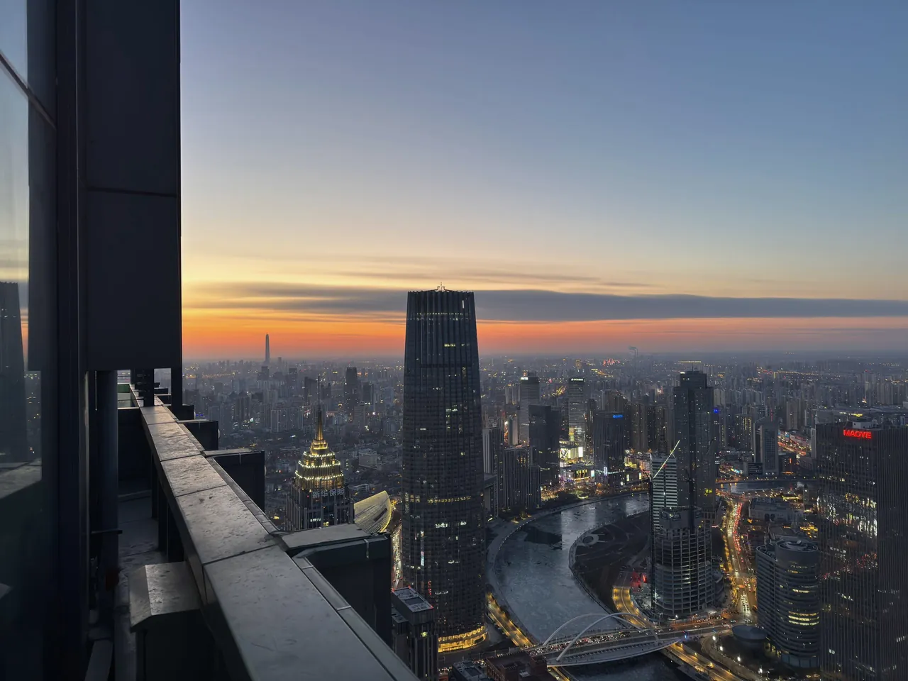 View from the edge of a high-floor balcony over Shanghai at dusk, lit towers and a curving river under an orange horizon.