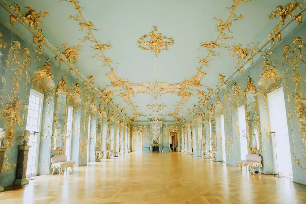 A long rococo gallery with pale green walls, gilded ornament, chandeliers and a polished parquet floor, empty.