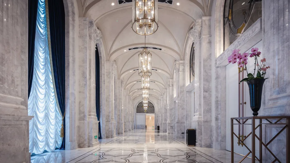 A long white marble corridor with vaulted ceiling, crystal chandeliers, blue curtains and an orchid arrangement.