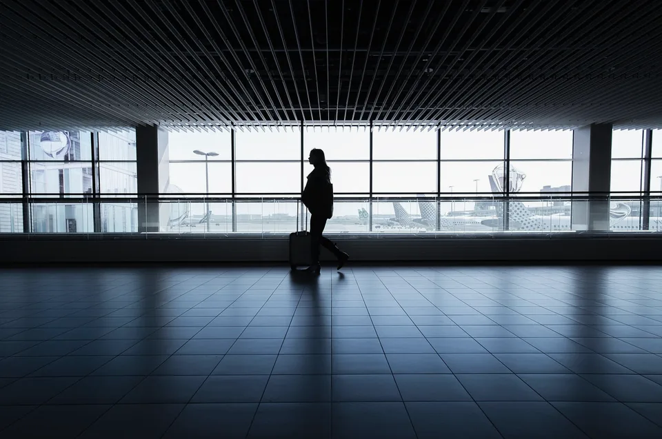 A lone traveller pulling a suitcase past a long wall of terminal windows, silhouetted against the light.