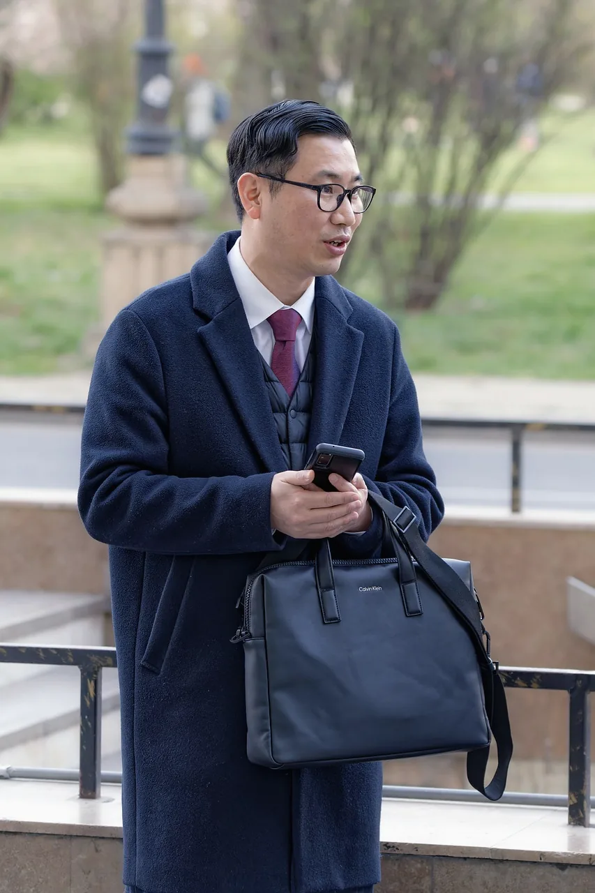 An East Asian man in glasses and a navy overcoat with a leather briefcase, holding a phone and looking off to the side outdoors.