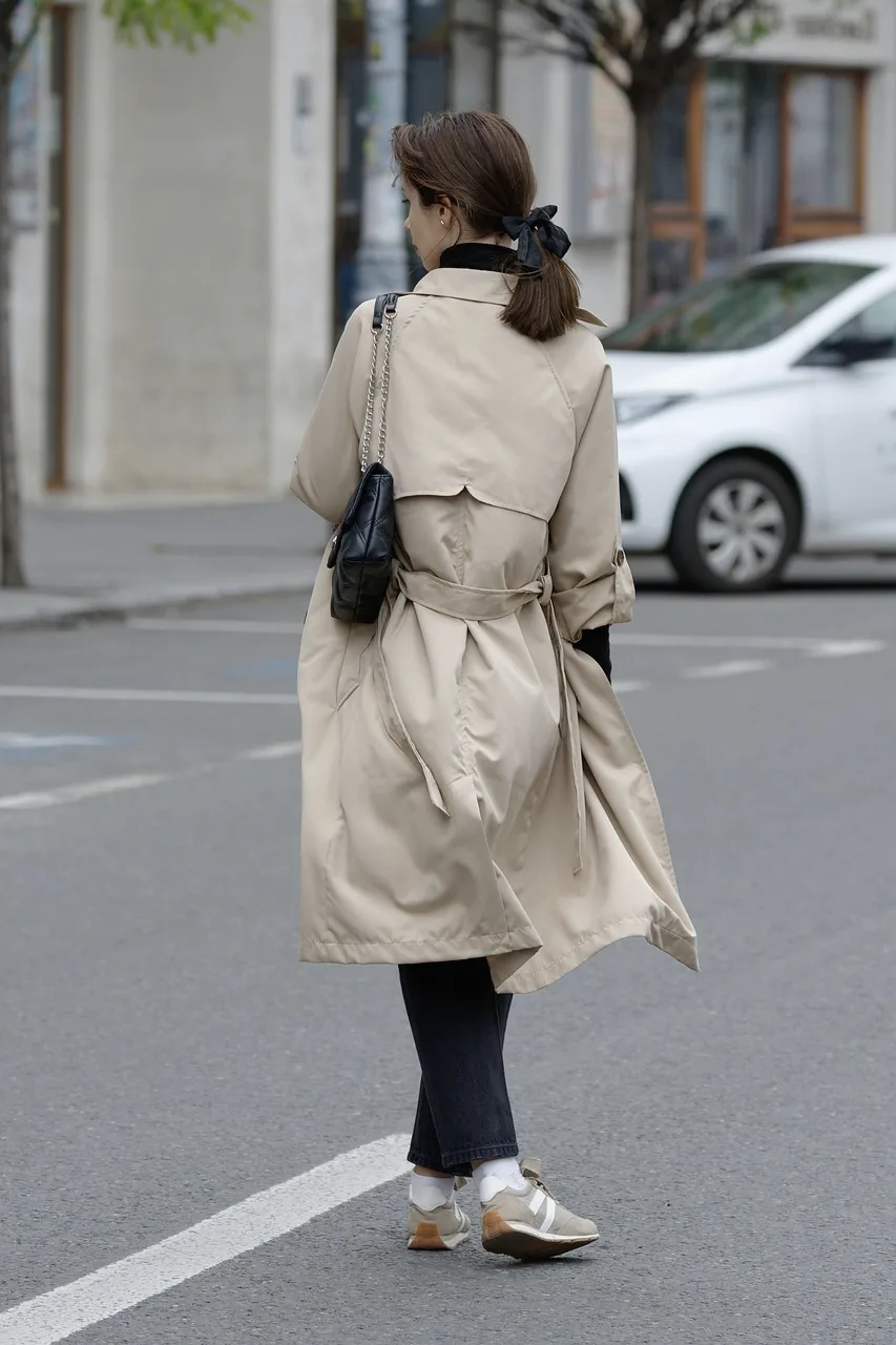 A woman in a beige trench coat walking away from the camera across a city street.