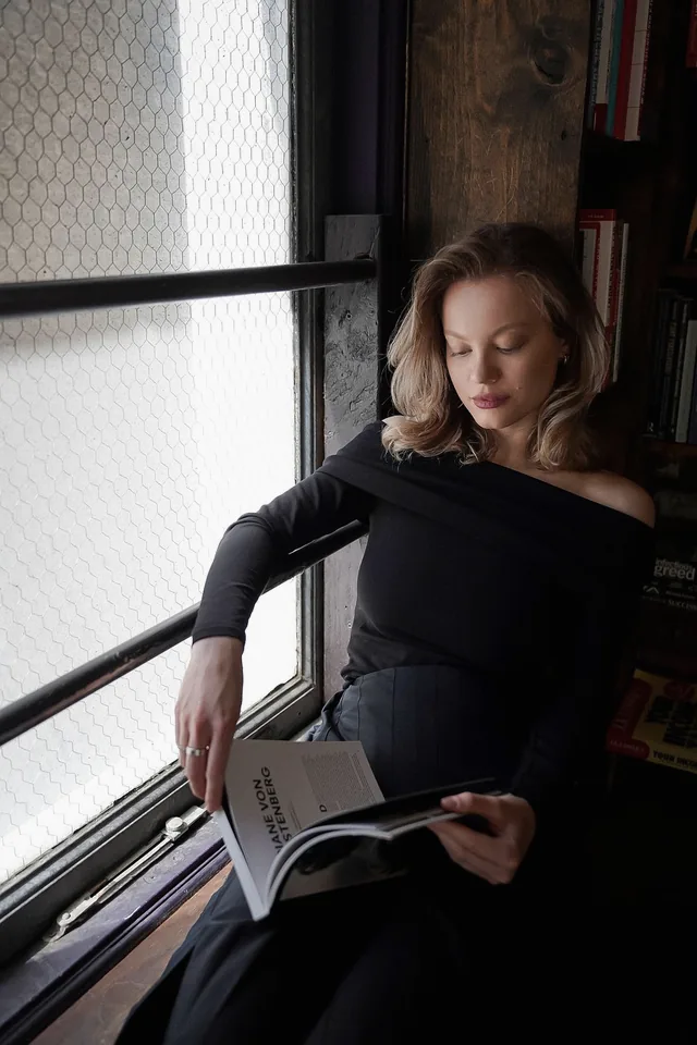 A woman in a black off-shoulder top reading a magazine by a tall window in a dim room.