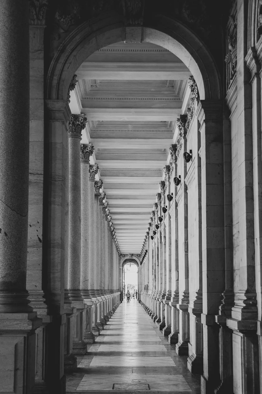 Black-and-white perspective down a long classical colonnade, stone columns and coffered ceiling receding to a bright end.