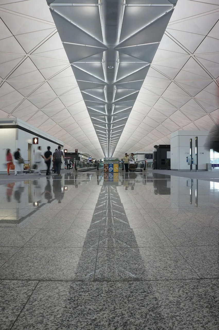A long airport terminal corridor with a white ribbed ceiling and polished stone floor reflecting it, a few travellers far away.