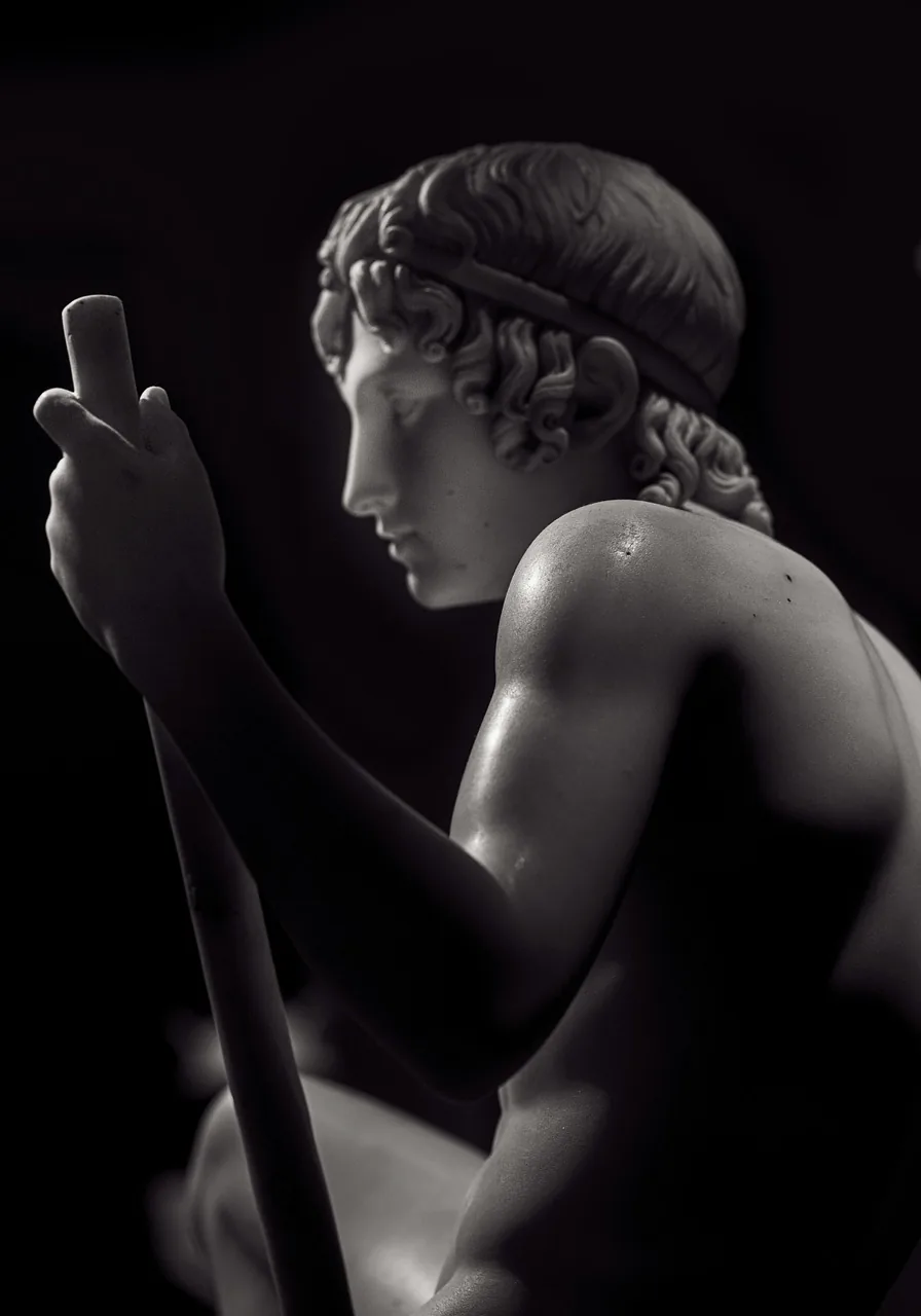 Marble sculpture of a seated young man in profile holding a staff, lit against a black background.