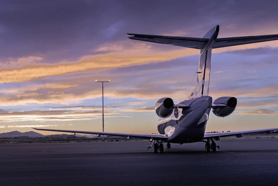 A small twin-engine business jet parked on an airport apron at dusk, seen from behind, under a violet and orange sky.
