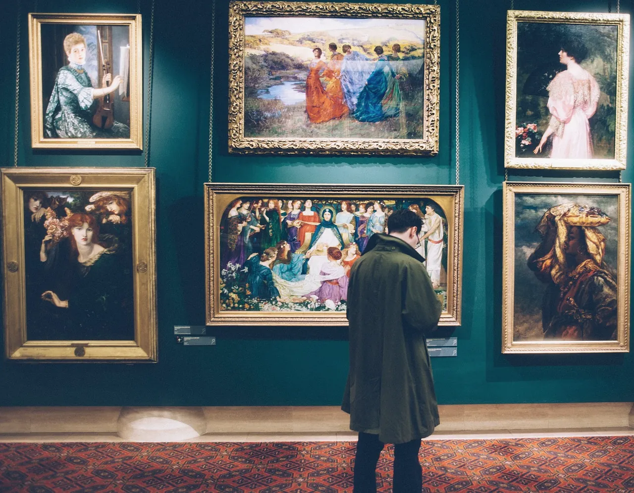 A man in a long green coat, seen from behind, studying a wall of framed Pre-Raphaelite paintings on a deep teal wall.