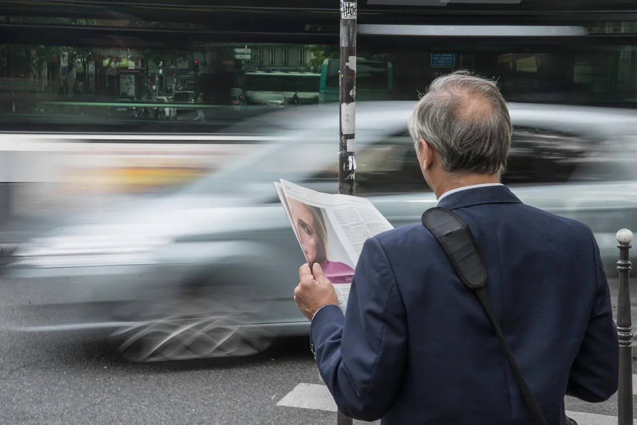 A grey-haired man in a navy blazer, seen from behind, reading a newspaper on a city pavement while a car passes in a motion blur.